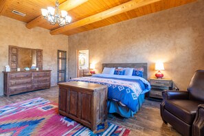 Une chambre avec un plafond en bois, un grand lit avec une literie bleue, une commode en bois, un fauteuil en cuir et un tapis coloré.