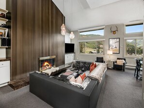 A modern living room with a fireplace, a sofa, a TV, and large windows offering a view of the outdoors. - Vantage Point Condo 601 (Vail)