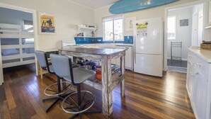 A kitchen with a wooden table, bar stools, a blue surfboard on the wall, and a refrigerator with a bird rock memorial.