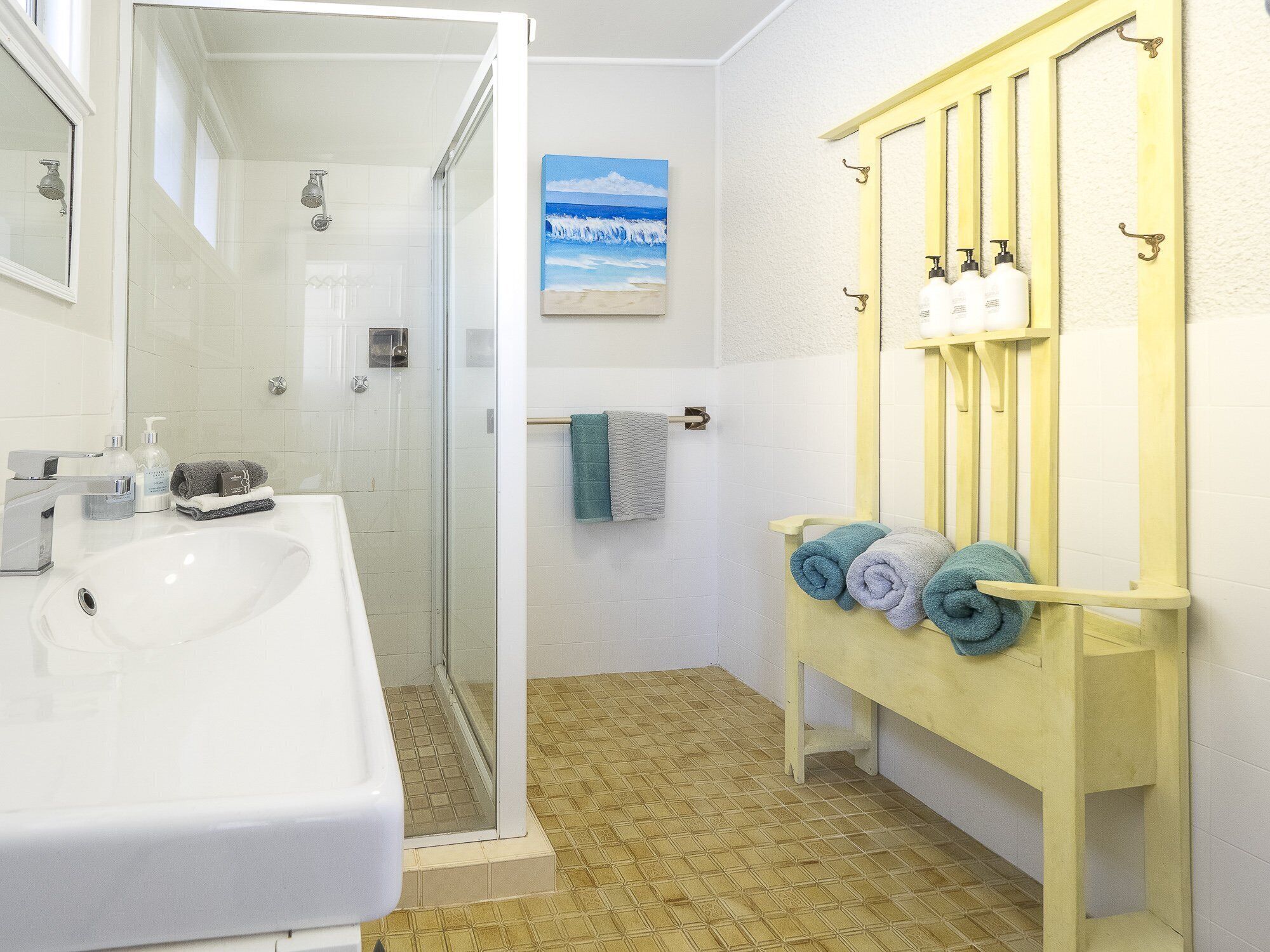 Une salle de bain avec un lavabo blanc, une cabine de douche en verre, un meuble-lavabo jaune avec un miroir et un porte-serviettes avec des serviettes.