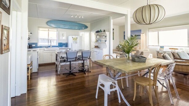 A spacious kitchen with wooden flooring, a dining area with a large table and chairs, and a living room with a sofa and a hanging light fixture.