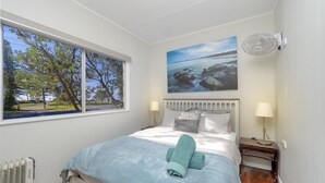 A bedroom with a large window, a bed with a blue blanket, a painting of a coastal scene, and a wall-mounted fan.