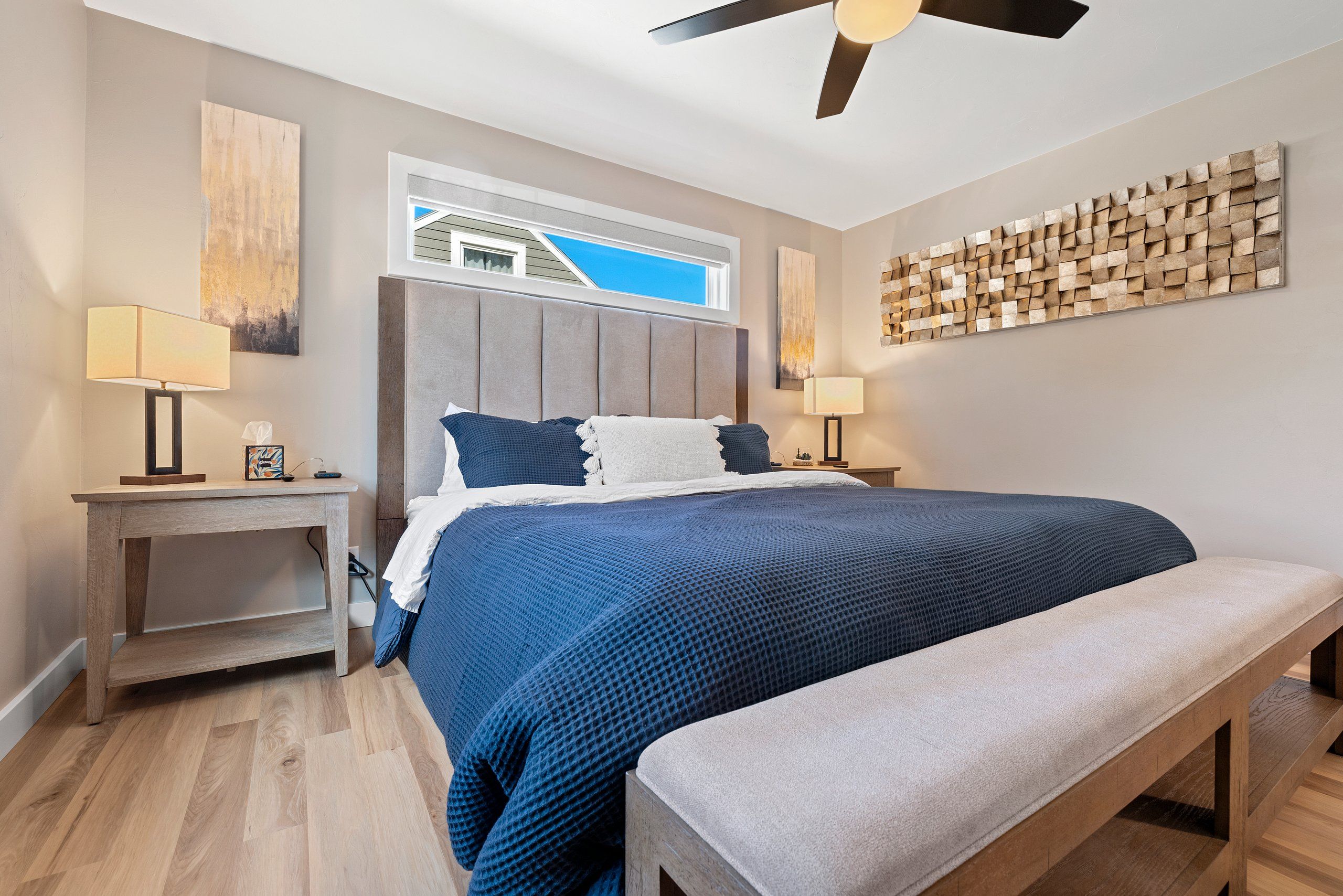 Un dormitorio con cama, mesitas de noche, un banco, ventilador de techo y obras de arte en las paredes.