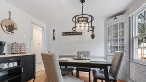 Een eetruimte met een houten tafel, vier stoffeerde grijze stoelen, een zwarte kast met keukengerei, een hanglamp en een raam met uitzicht.