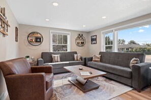 A spacious living room with a leather sofa, a grey couch, and a coffee table. There are two windows providing natural light and a view of the outside.