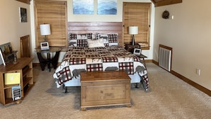Une chambre à coucher avec un grand lit, une tête de lit en bois, deux fenêtres équipées de stores, une table de chevet avec des lampes, une petite commode en bois et un sol recouvert d’une moquette.