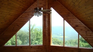 Une pièce mansardée en bois, avec un plafond en pente et un ventilateur de plafond. De grandes fenêtres offrent une vue sur un paysage boisé.