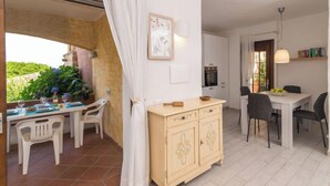A dining area with a table and chairs, a kitchen with cabinets, and a view of greenery outside. - Apartment  Trudi by Interhome (Cannigione)