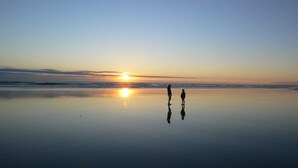 Eine Strandansicht, zwei Personen stehen im Sand und blicken bei Sonnenuntergang auf das Meer.