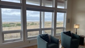 Ein Zimmer mit großen Fenstern, die auf einen Strand Blick bieten, zwei smaragdgrüne Sessel, eine Lampe und ein gemustertes Teppich.