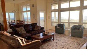 Ein geräumiges Wohnzimmer mit einem braunen Ledersofa, einem blauen Sessel und einem Couchtisch. Große Fenster bieten einen Ausblick ins Freie.