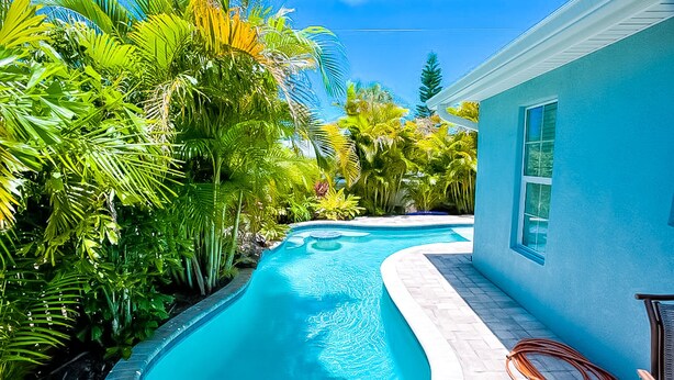 Une piscine entourée d’une végétation tropicale luxuriante, un bâtiment bleu et un ciel d’un bleu limpide.