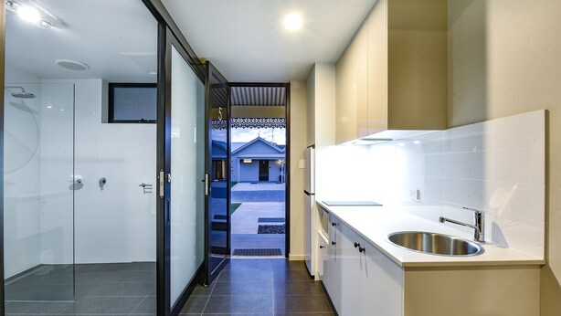A modern bathroom with a glass shower enclosure, a sink with a faucet, and a view of a house through an open door. - Aloha Central Premium Studios (Mount Gambier)