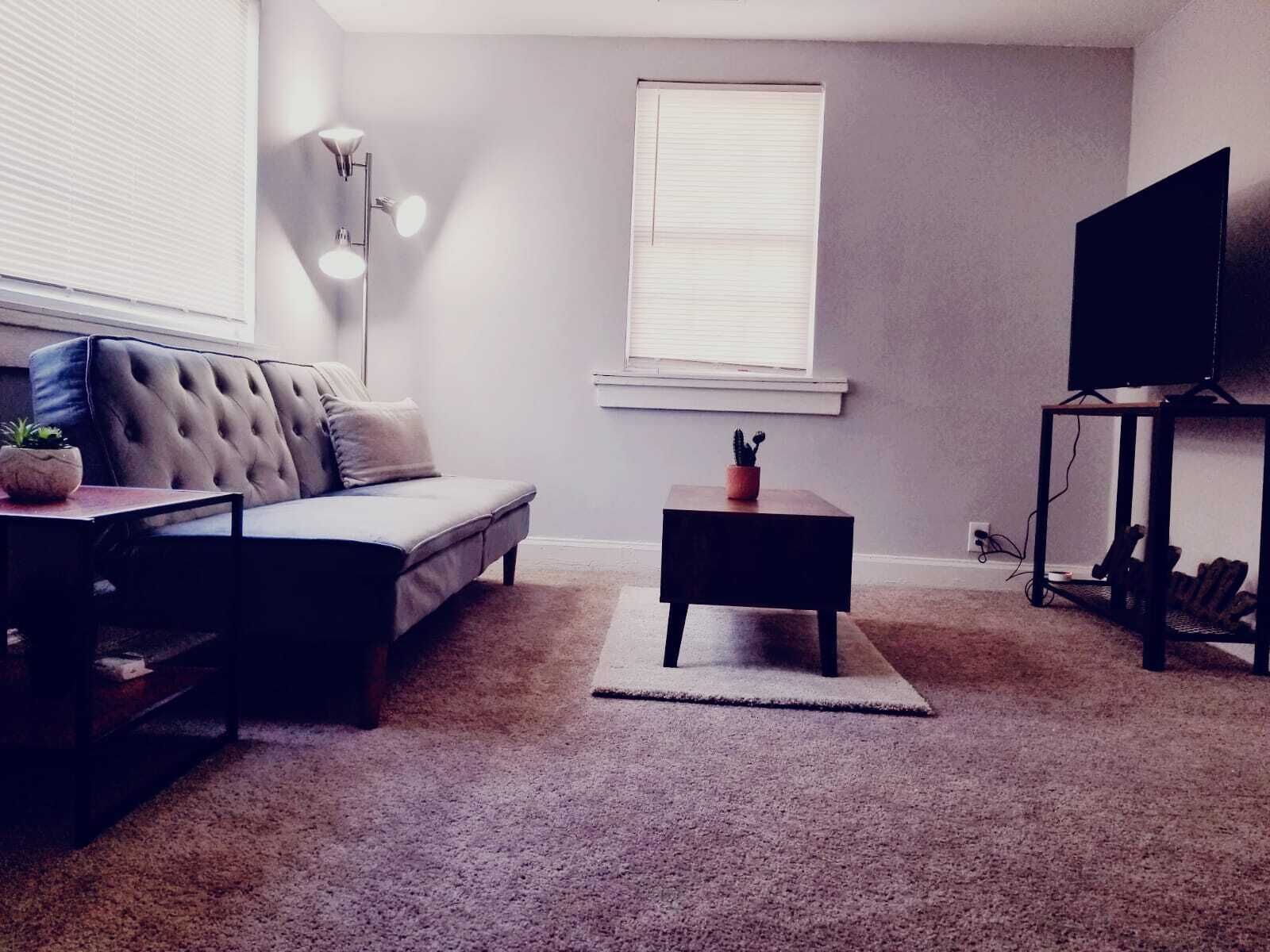 Una sala de estar con un sofá, una mesita, una lámpara de pie, un televisor sobre un mueble bajo y una ventana con persianas.