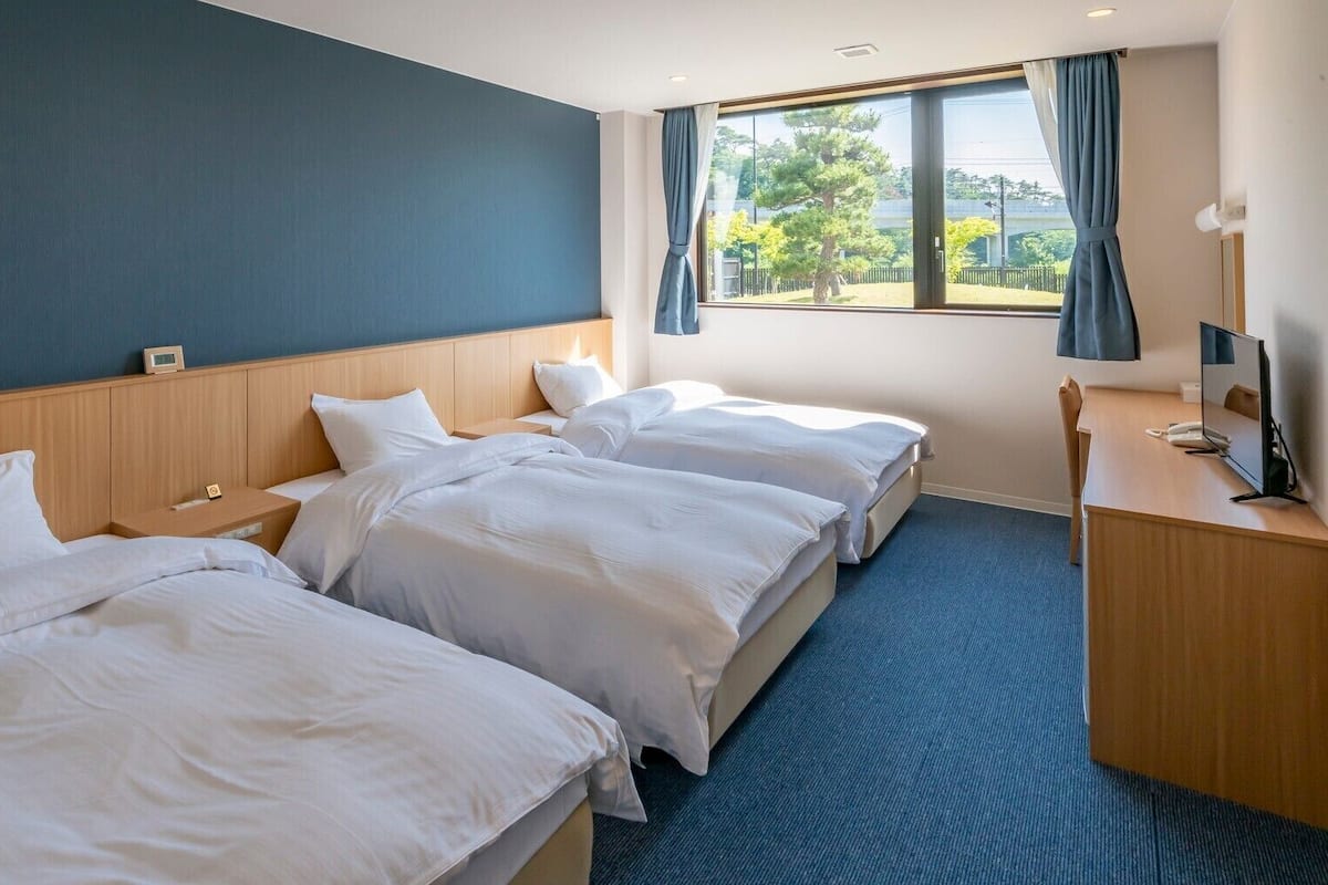 Quarto de hotel com duas camas, uma mesa, uma televisão e uma janela com cortinas.
