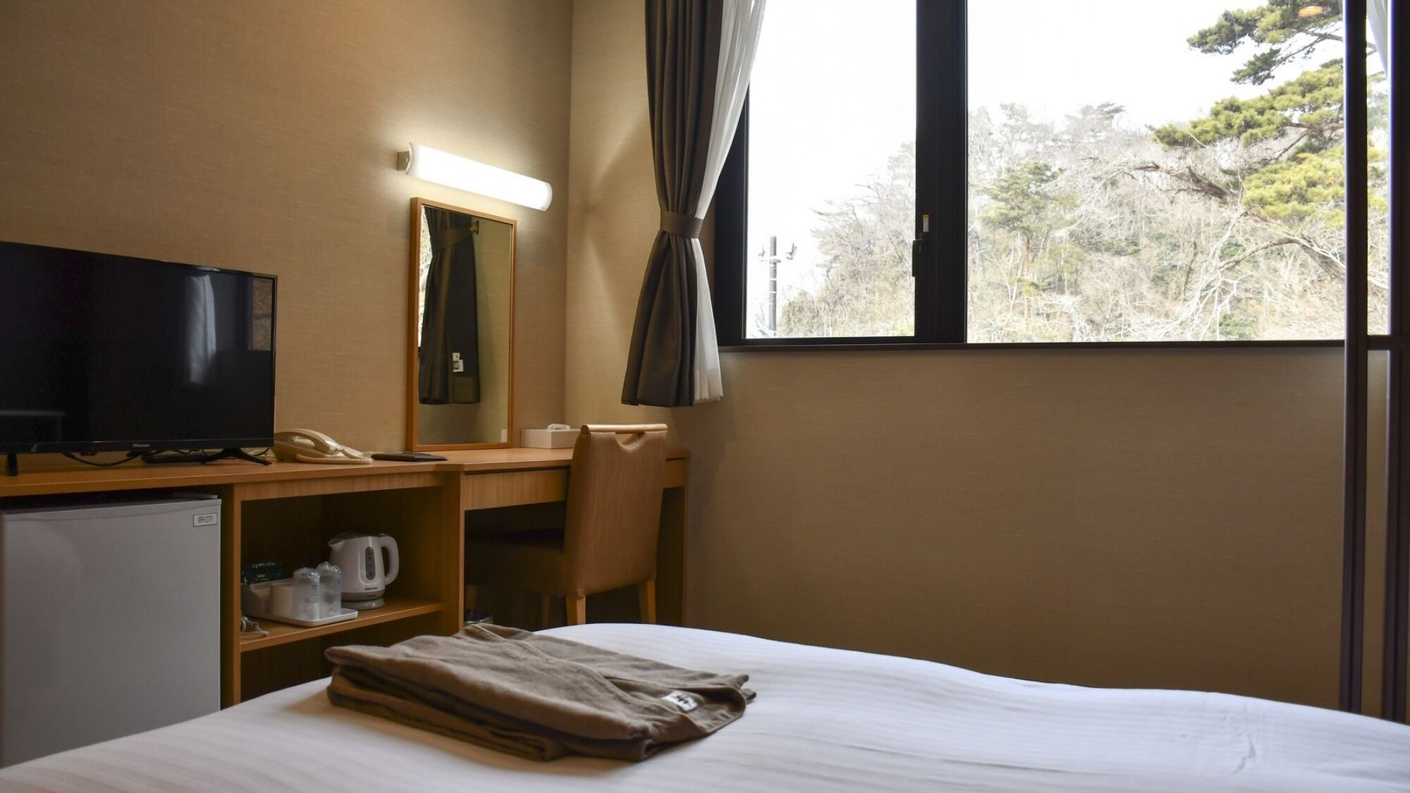 A hotel room with a bed, a desk with a chair, a television, a mirror, a window with curtains, and a view of trees outside.