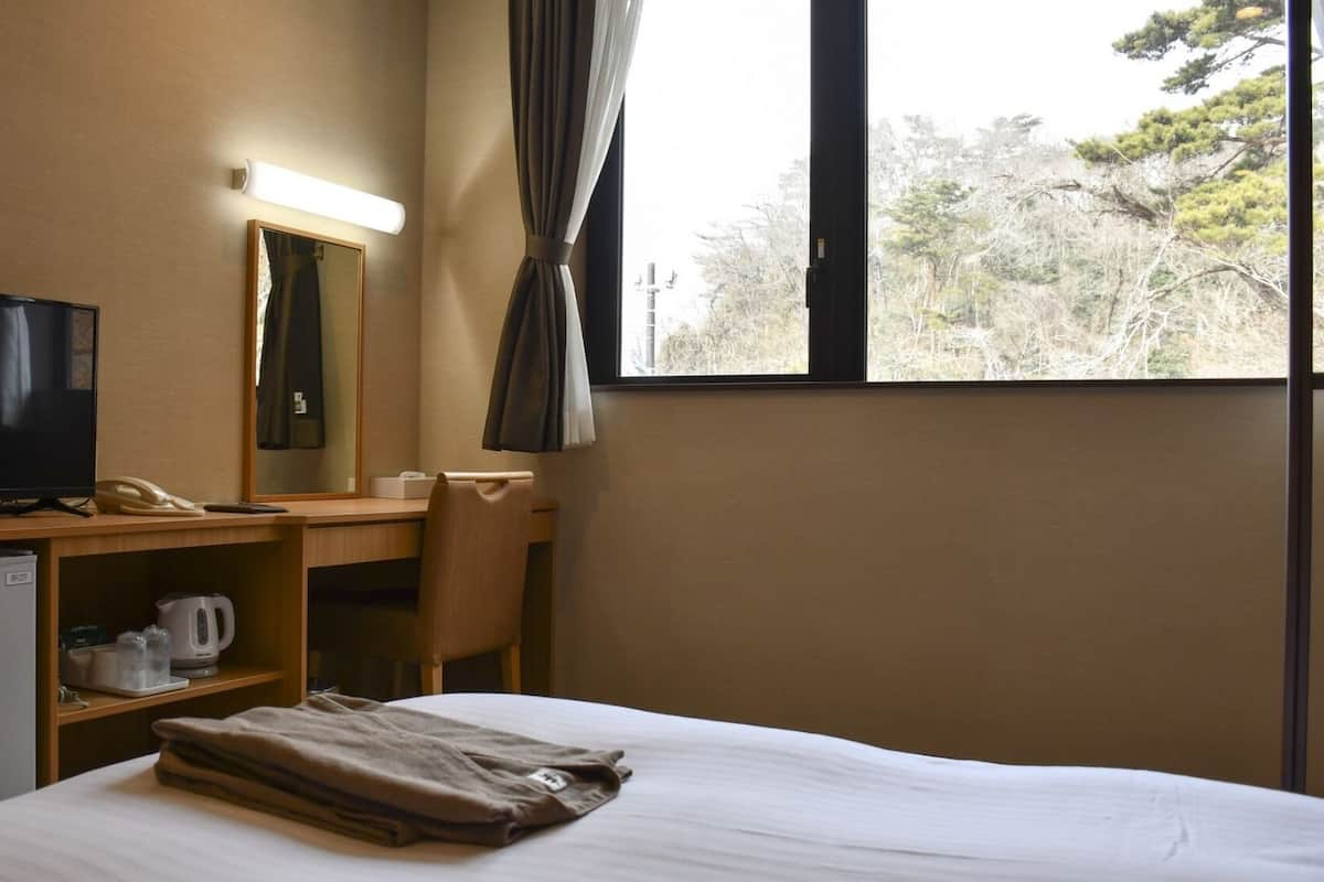 Quarto de hotel com cama, escrivaninha com cadeira, televisão, espelho, janela com cortinas e vista para árvores.