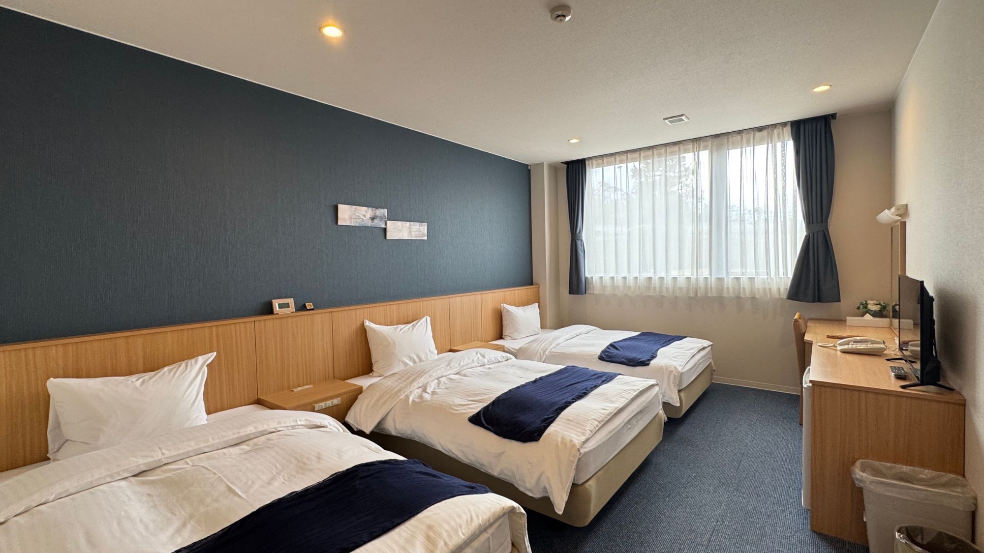 A hotel room with three beds, a desk, a chair, a trash bin, and a window with curtains.