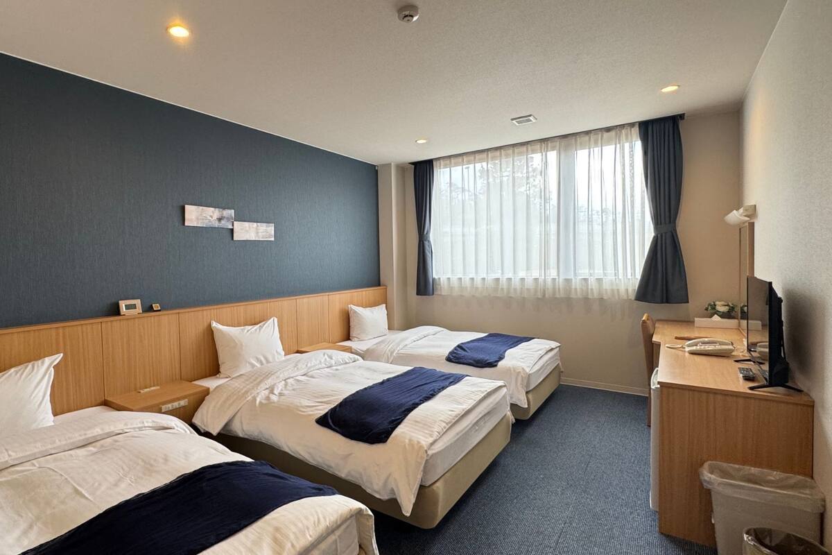 Quarto de hotel com três camas, uma escrivaninha, uma cadeira, um cesto de lixo e uma janela com cortinas.