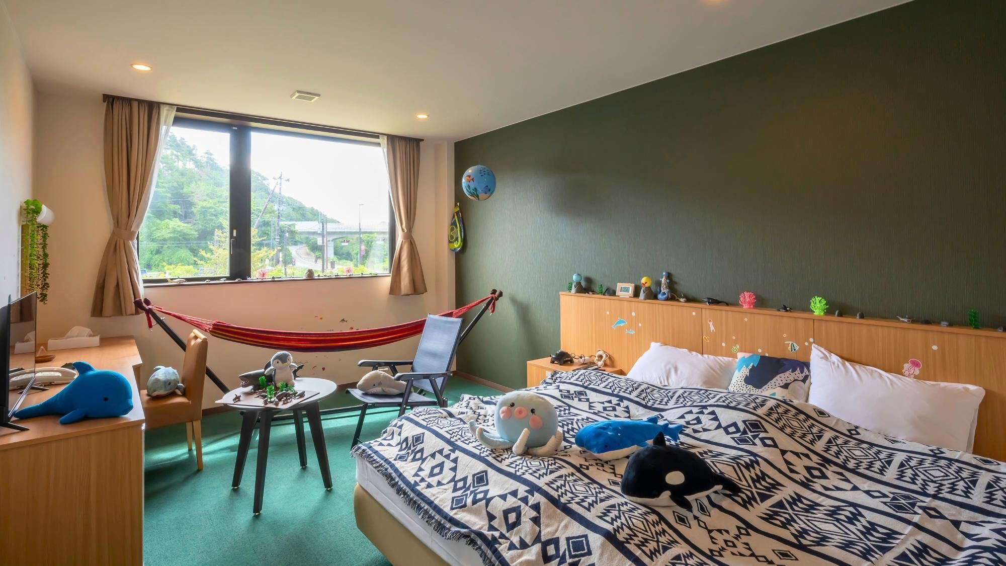 A bedroom with a bed, a desk, a chair, a hammock, and a view of greenery outside.