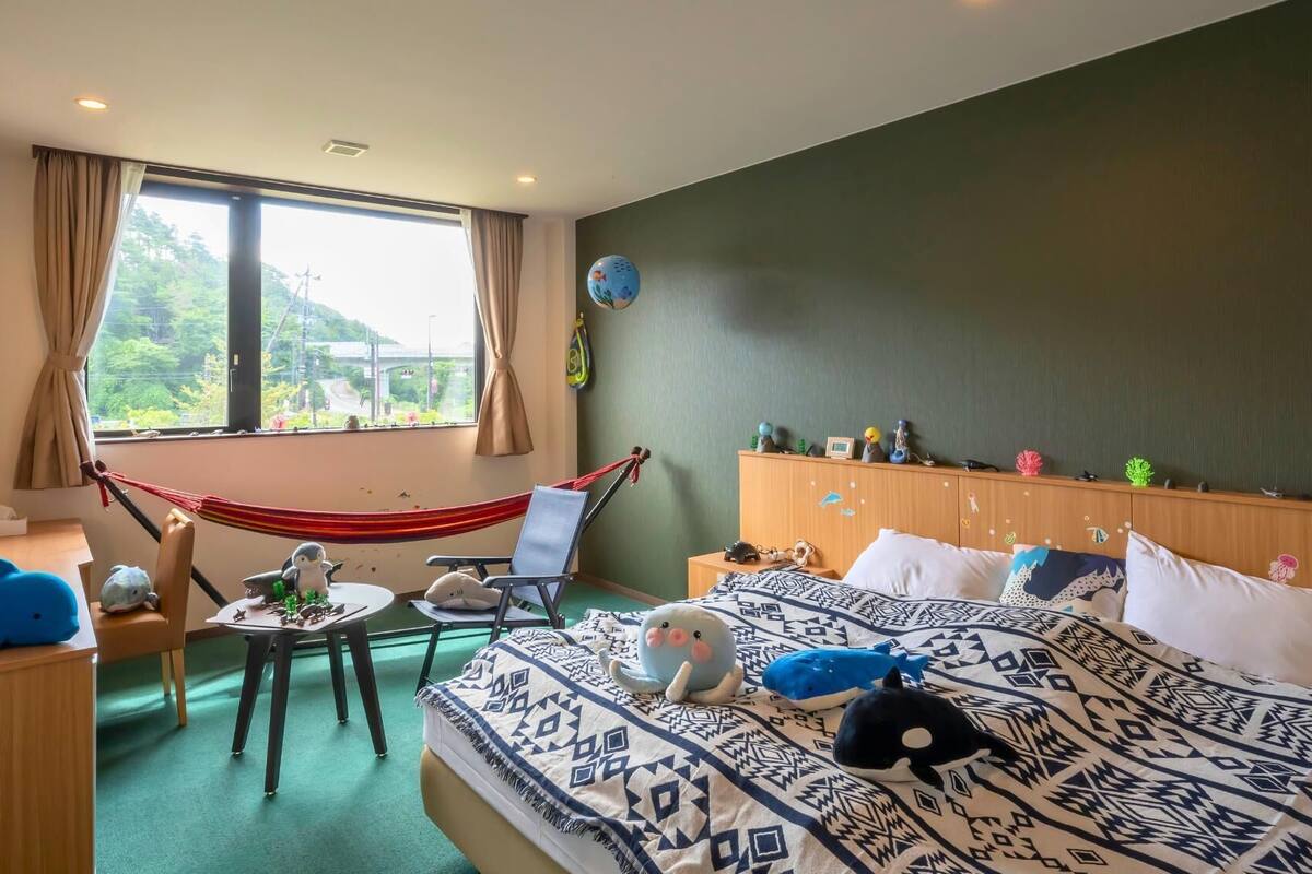 Quarto com cama, escrivaninha, cadeira, rede e vista para o verde lá fora.