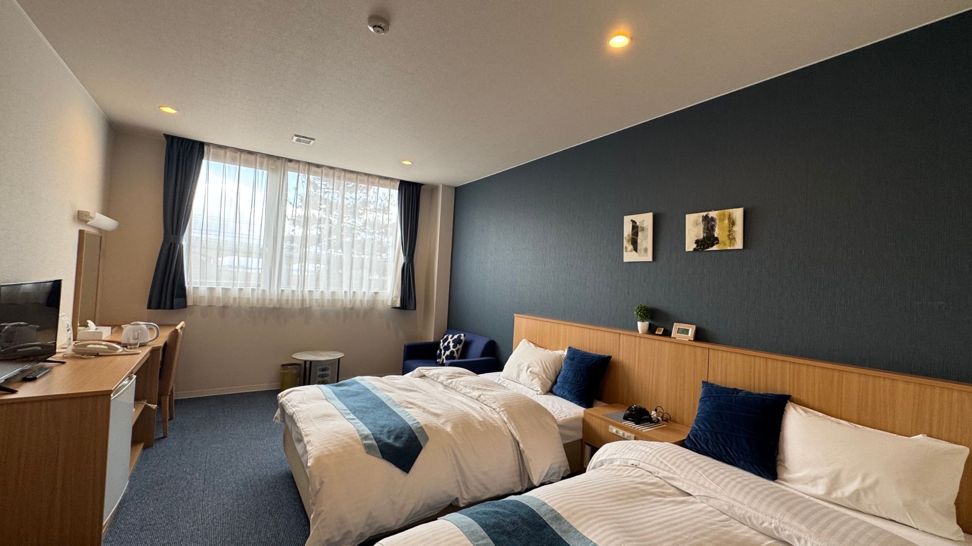 A hotel room with two beds, a desk, a chair, a small table, and a window with curtains.