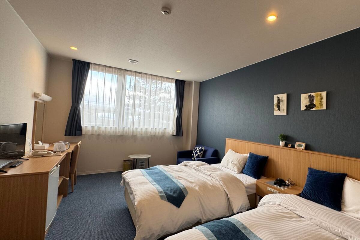 Quarto de hotel com duas camas, uma escrivaninha, uma cadeira, uma mesinha e uma janela com cortinas.