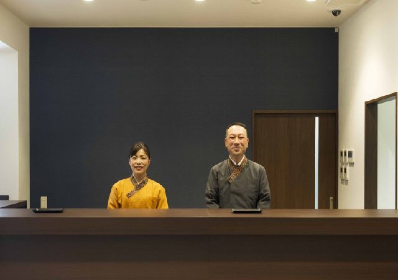 A reception desk with two individuals standing behind it, a dark wall, and a wooden door.