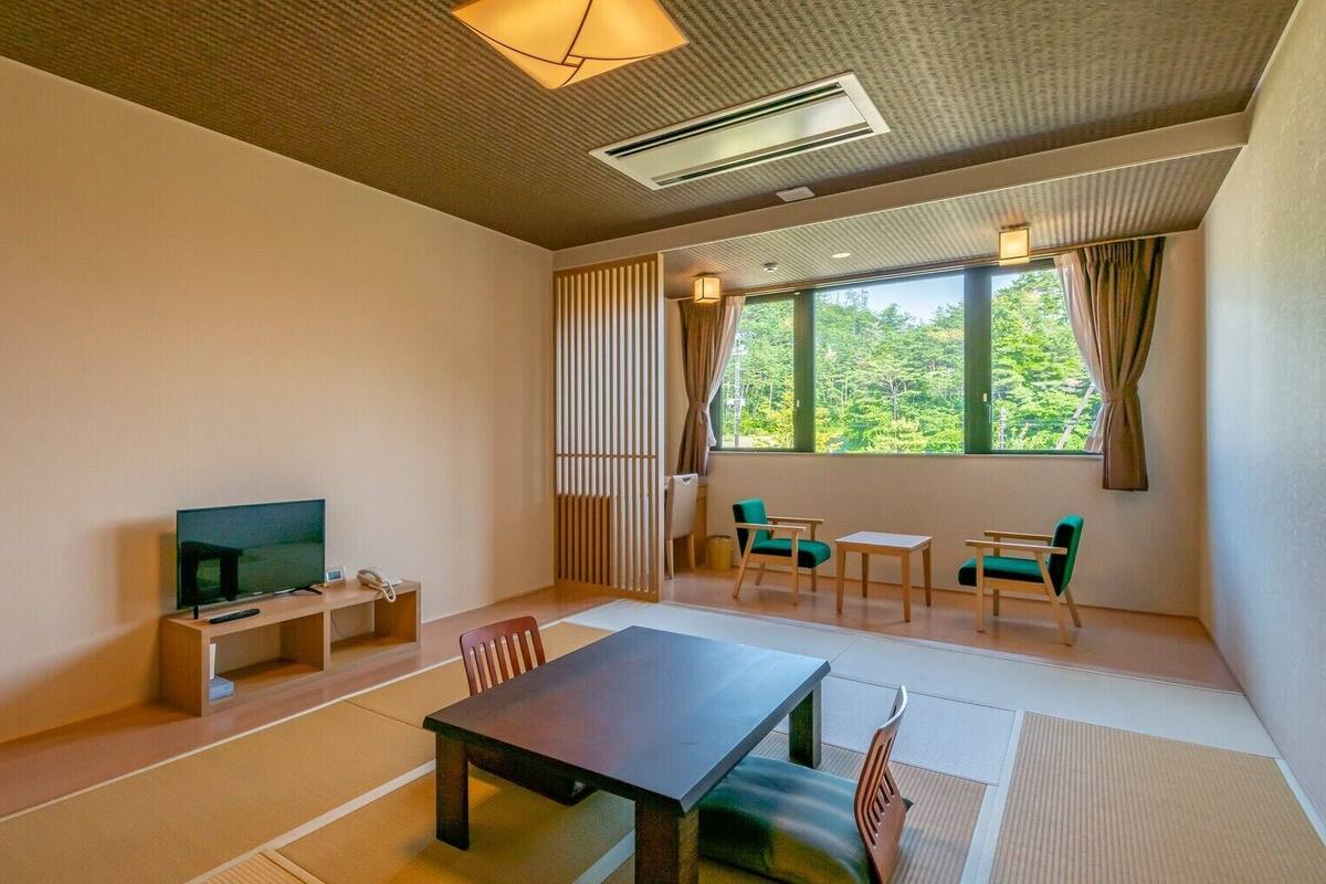 Quarto com televisão, mesa, cadeiras, janela com vista para vegetação e teto com iluminação embutida.