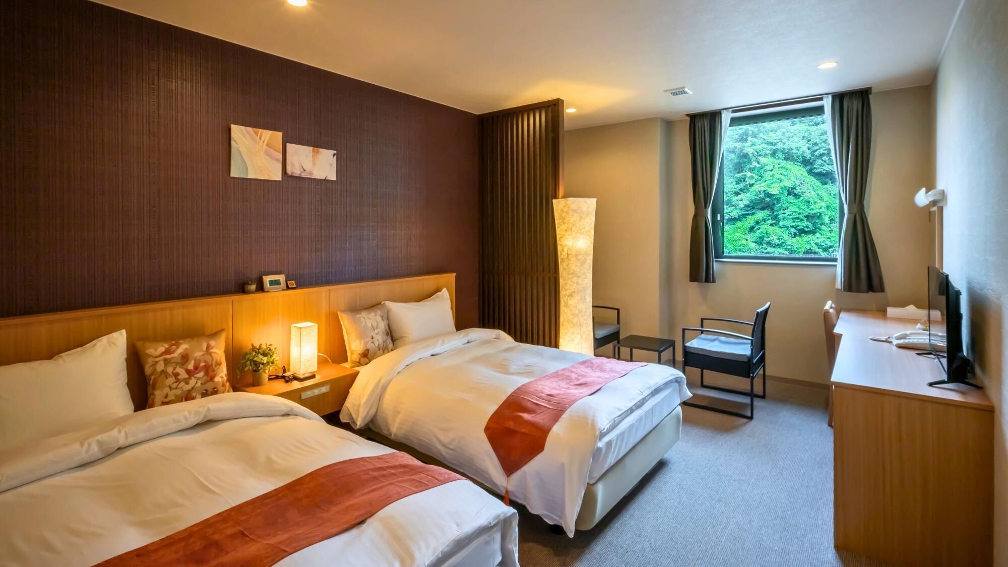 A hotel room with two beds, a desk, a chair, a TV, and a window with a view of greenery.