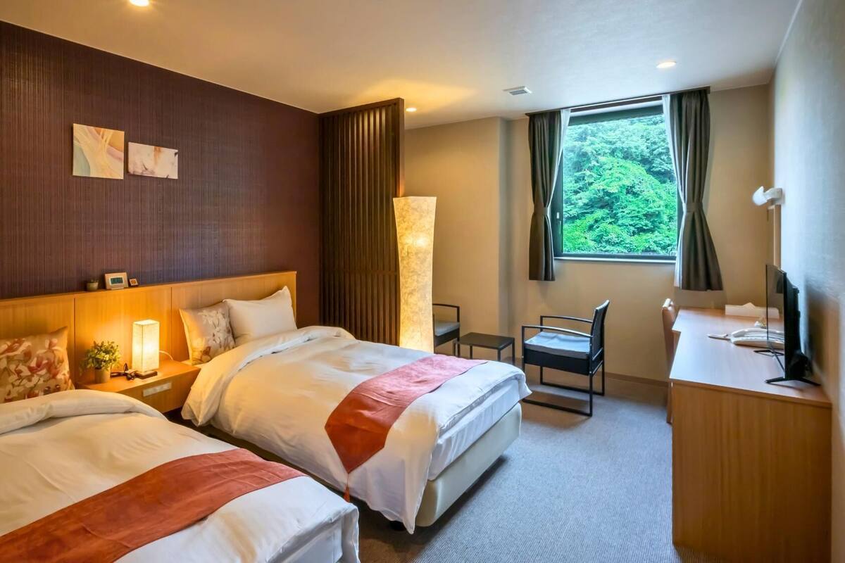 Quarto de hotel com duas camas, uma escrivaninha, uma cadeira, uma TV e uma janela com vista para o verde.