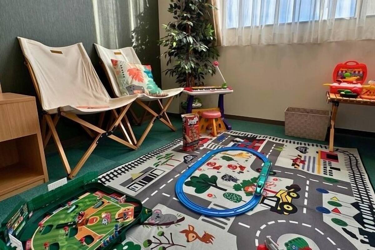 Uma sala de brinquedos com piso coberto de carpete, cadeiras, um conjunto de trens de brinquedo e uma mesa pequena com brinquedos.