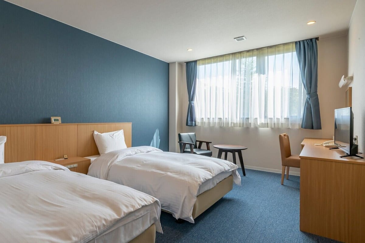 Quarto de hotel com duas camas, uma escrivaninha, uma cadeira, uma televisão e uma janela com cortinas.