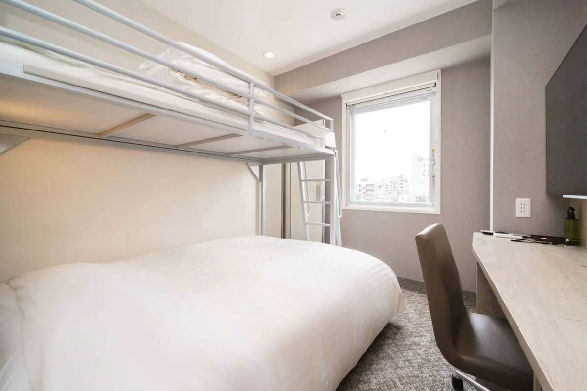 A hotel room with a bunk bed, a desk, and a window with a city view.