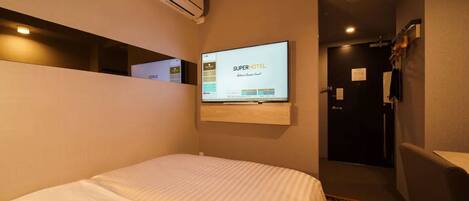 A hotel room with a bed, a mounted TV displaying the hotel's name, a coat rack, and a door.