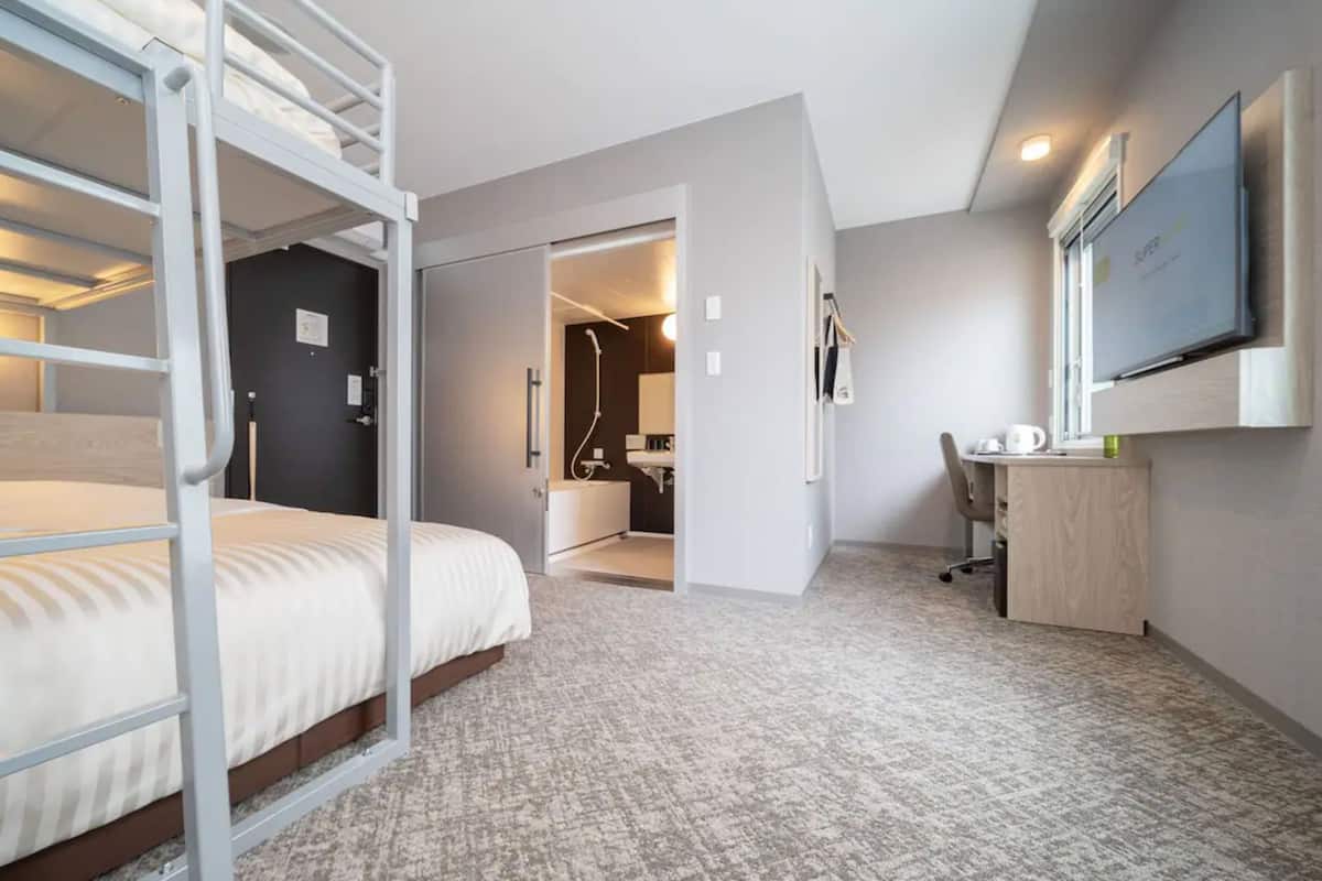 A hotel room with a bunk bed, a desk, a chair, a TV, and a bathroom visible through an open door.