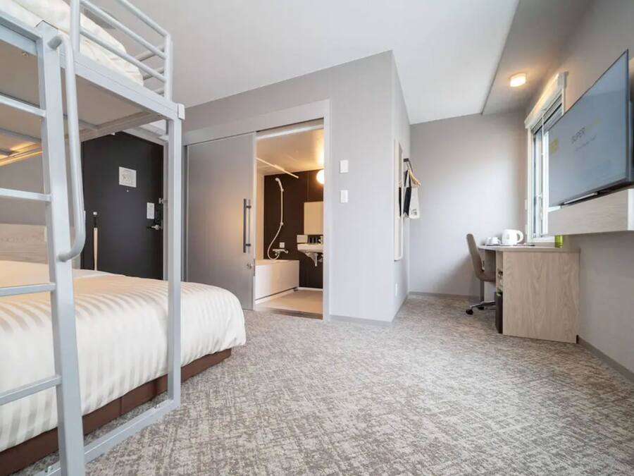 Ett hotellrum med en våningssäng, ett skrivbord, en stol, en tv och ett badrum som syns genom en öppen dörr.