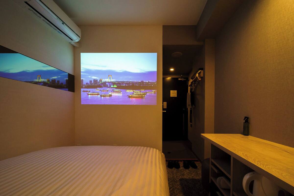 A hotel room with a bed, a small desk, a shelf with toiletries, and a wall-mounted projector displaying a cityscape.