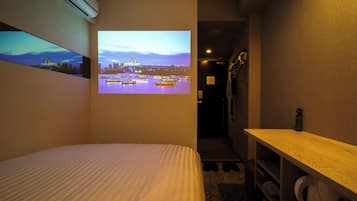 A hotel room with a bed, a small desk, a shelf with toiletries, and a wall-mounted projector displaying a cityscape.