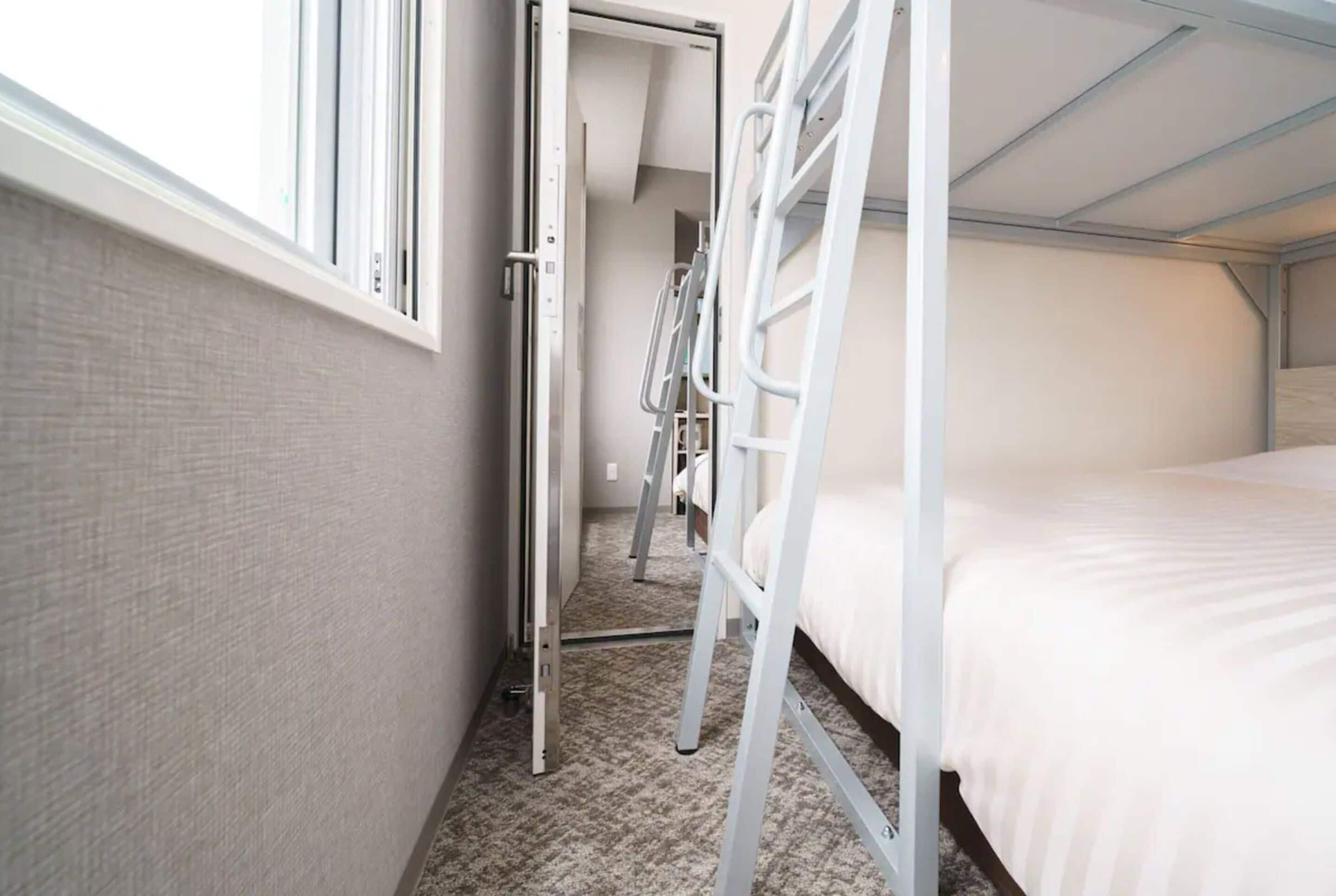 Et smalt hotelværelse med køjesenge, et vindue og en dør, der fører til en gang.