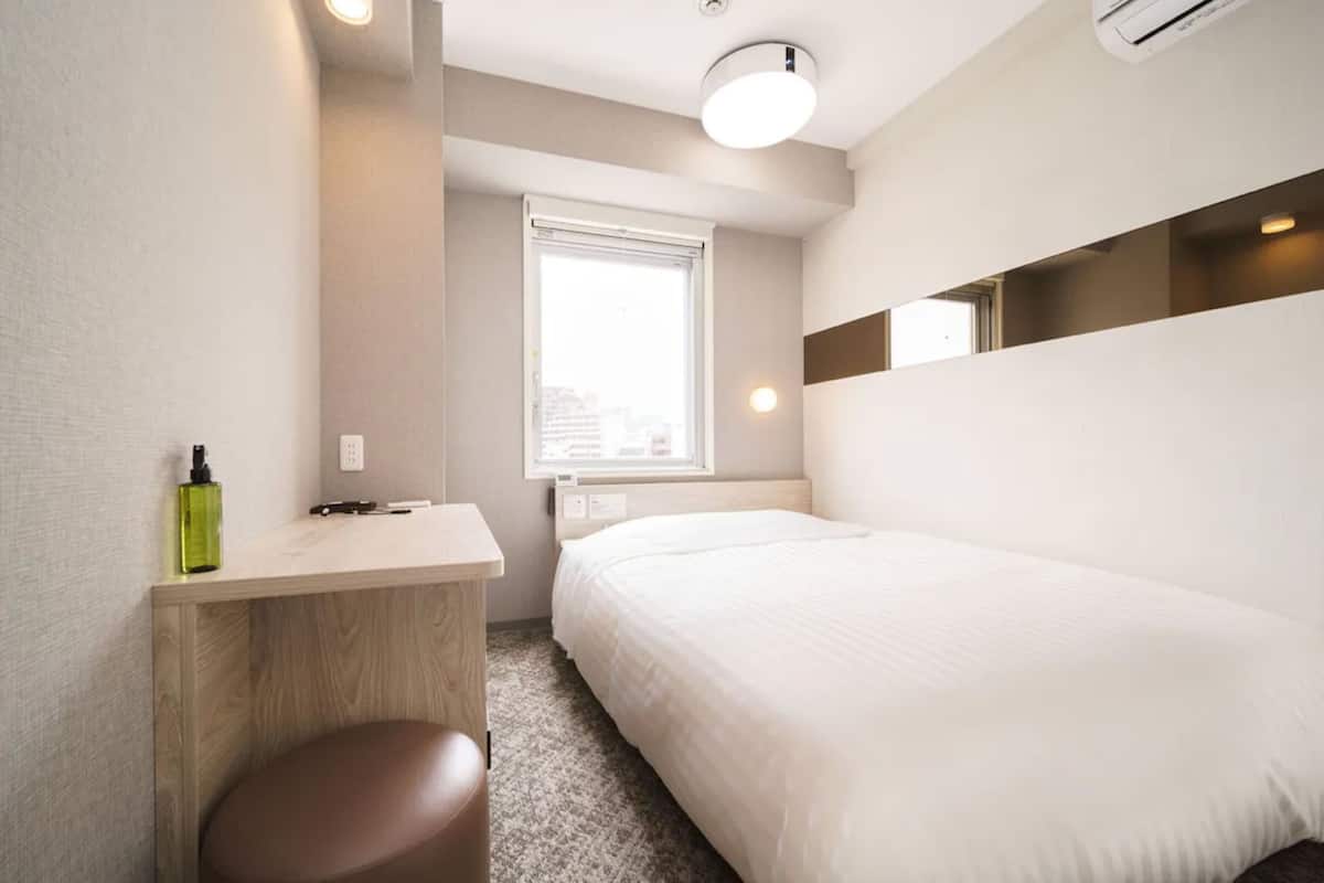 A modern hotel room with a bed, a small desk, a chair, and a window with a city view.