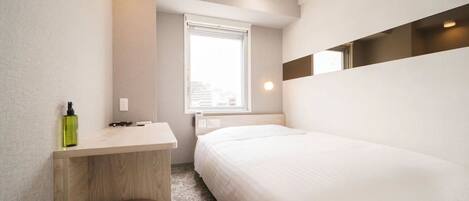 A modern hotel room with a bed, a small desk, a chair, and a window with a city view.