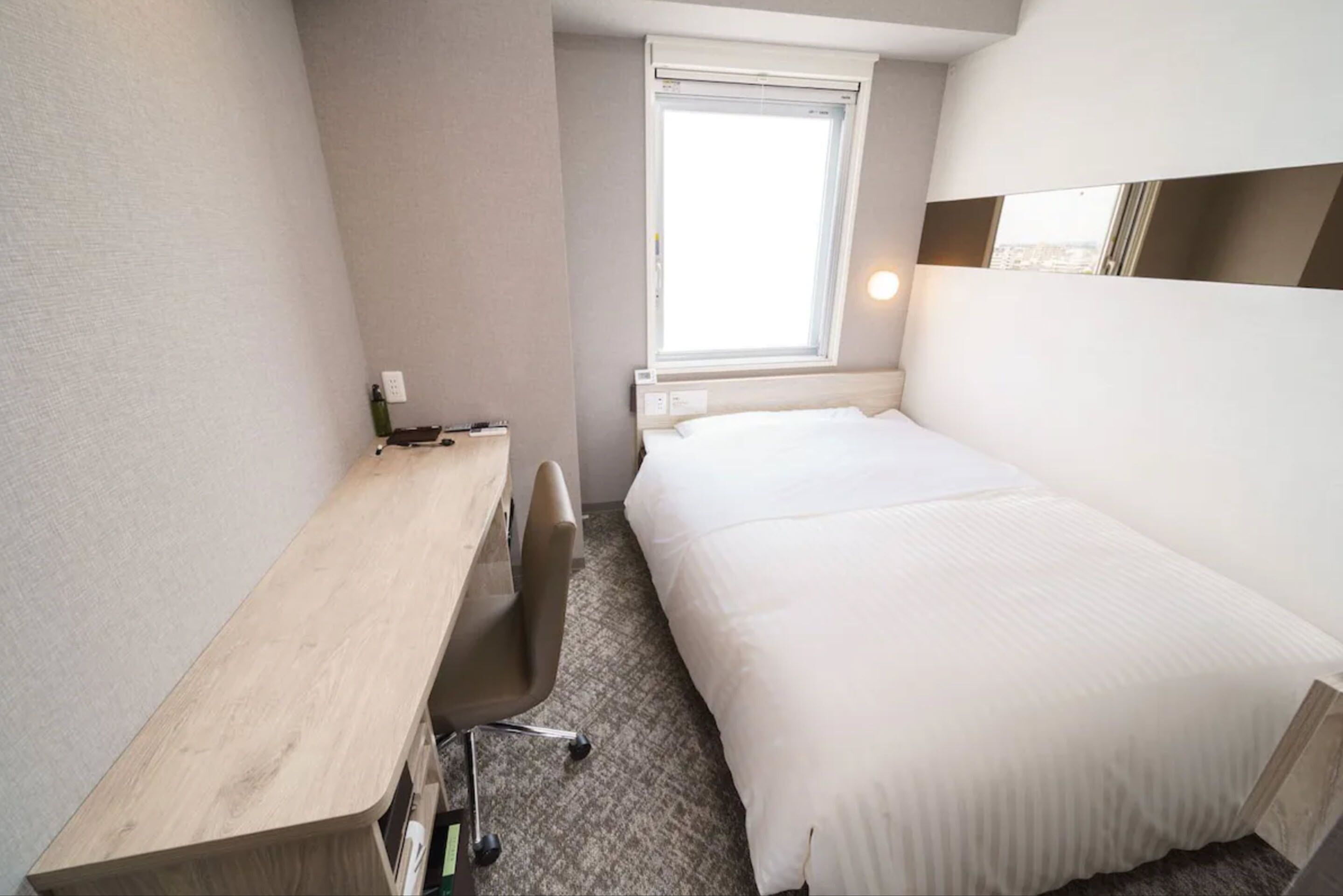 Een hotelkamer met een bed, een bureau, een stoel, een raam en een wandlamp.