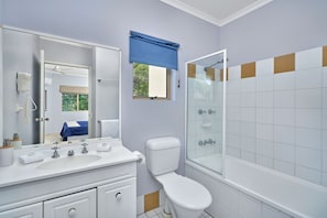 Une salle de bain avec un lavabo blanc, un miroir et des toilettes, une douche avec une porte en verre et une fenêtre avec des stores bleus.