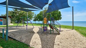 A playground with sand-covered ground, a slide, and a swing set, surrounded by trees and a view of the sea. - Tropical Oasis near Beach (Holloways Beach)