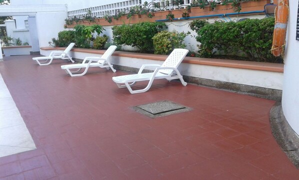 A row of white sun loungers on a red-tiled patio with a white wall and green plants in the background.