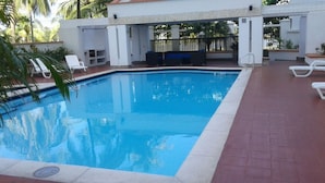 Piscina retangular com borda azulejada, rodeada por um pátio de tijolos. Há cadeiras de descanso brancas e um sofá azul perto da piscina.