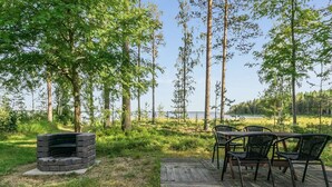 En utomhusplats med sittmöbler, en eldstad av sten och metallstolar, omgiven av höga träd och med utsikt över en sjö.