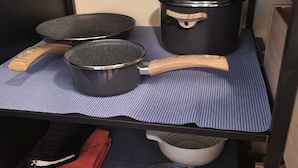 Un coin cuisine avec un tapis à carreaux bleus, deux casseroles noires à manches en bois, une passoire blanche, un saladier en verre et un panier tressé contenant des blocs de bois et un tissu rouge.