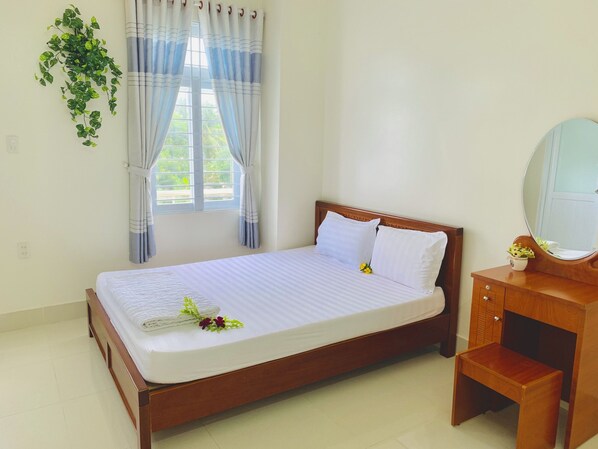 Una cama bien tendida con sábanas y almohadas blancas, una mesita de noche de madera con un espejo, y una planta en maceta colgando en la pared.